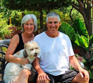 A man and woman sit outside on lawn furniture with their dog.