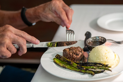 A plate with filet mignon, asparagus and mashed potatoes, with a hand with knife and fork poised above it.
