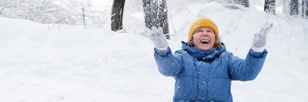 Winter scene of a beautiful smiling senior woman in a blue coat and white gloves with her hands raised toward falling snow.