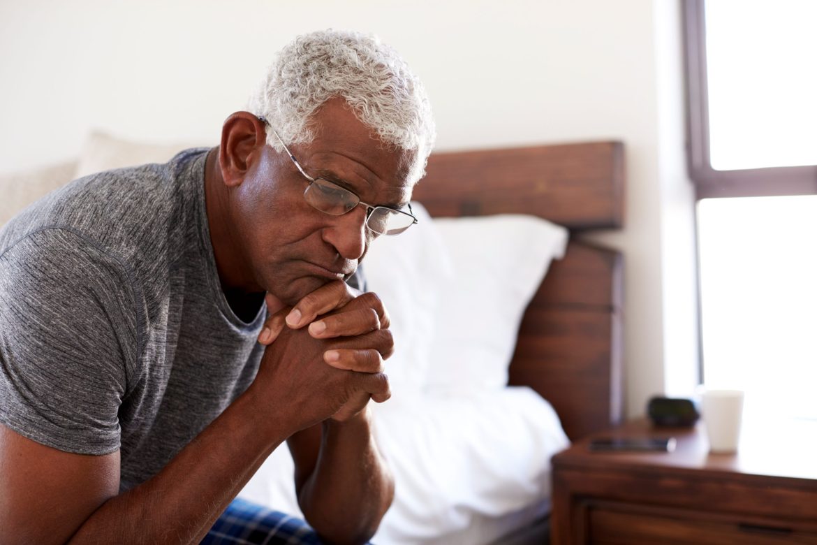 Older man looking worried sitting on a bed.