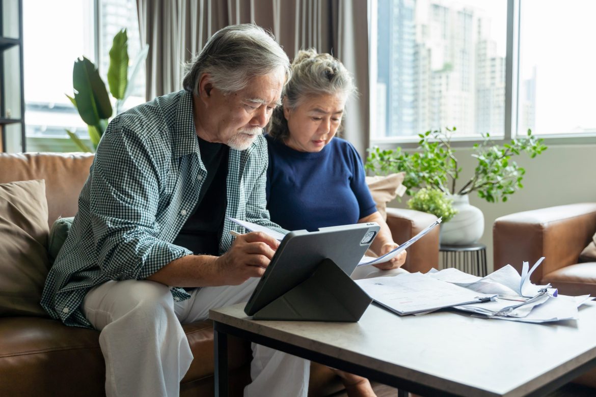 An elderly couple look at stack of papers and computer tablet as they plan for long-term care in a nursing home.