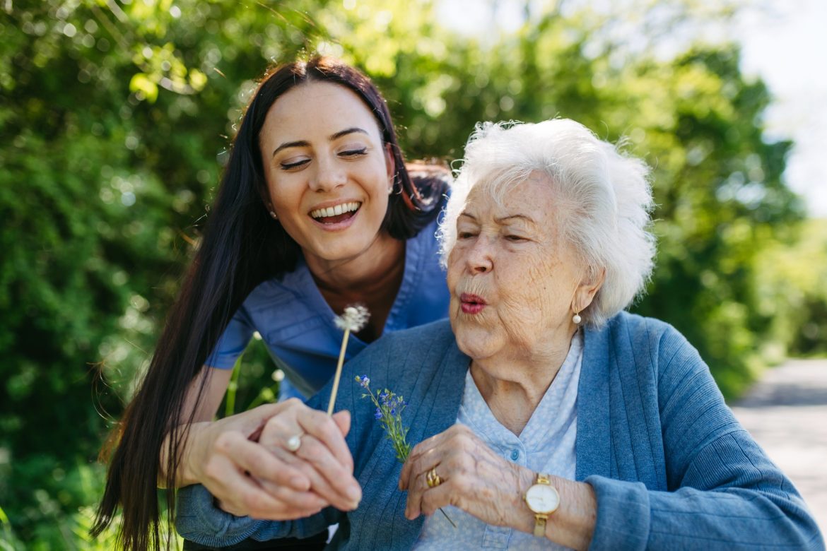 Older woman in a wheelchair with a caregiver helping her blow the fluff off a dandelion.