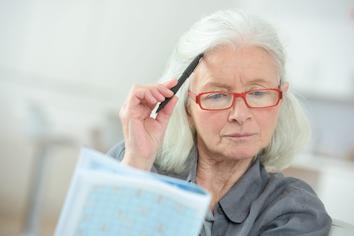 An older woman puts her pen to her head while struggling to remember something.