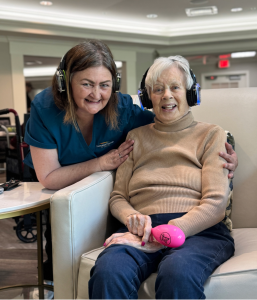 A memory care associate has her arms on the shoulders of a resident. Both are wearing the Disco Memory headphones.