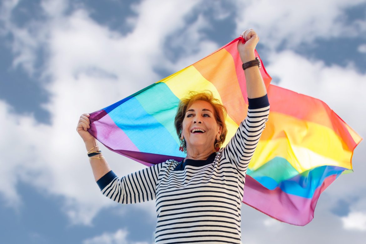 Older LGBTQ+ woman waving rainbow flag under blue sky